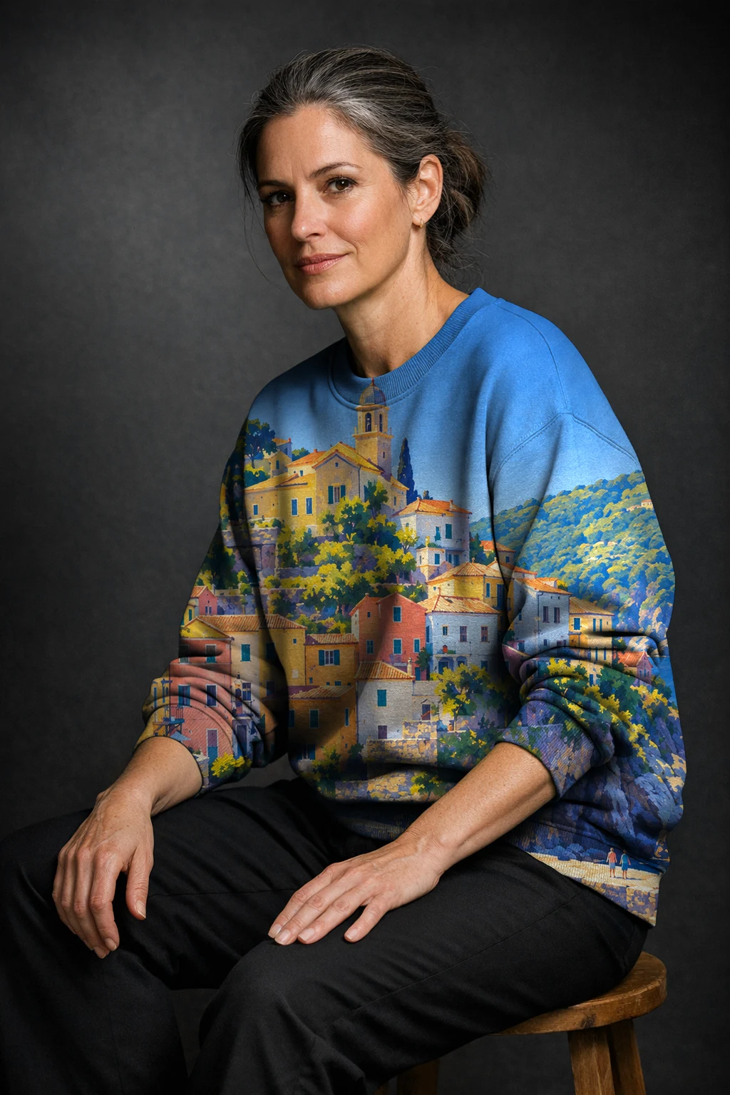 A woman seated on a stool in a sweatshirt printed edge to edge with the Riviera Gouache 1930 harbour